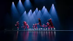 Dancers wear red suits and jump in the air. There is a line of white chairs in the background.