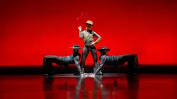 One dancer in a hat stands confidently in front of a deep red backdrop, flanked by 2 other dancers on either side.