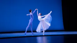 Two ballet dancers perform on stage. The ballerina holds a high arabesque, her tulle skirt sweeping around her feet, while her partner supports her at her side.