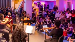 D-Composed playing in front of kids sitting on floor mats and an audience seated in the Harris Theater lobby.