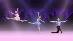 Three ballet dancers pose on stage before a purple city skyline; one leaps, one balances on pointe, and one kneels with arms extended.