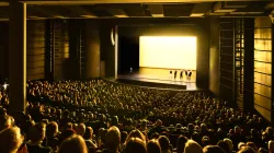 A full Harris Theater auditorium. The audience watches a dance performance where a yellow light is emitted.