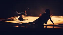A dancer resting on the stage floor while another dancer spins on their head on top of a large drape.