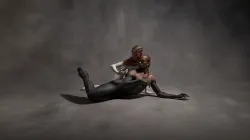 Two dancers lay on a gray backdrop. One dancer is covered in black paint up to their shoulders. The second dancer is covered in white paint.