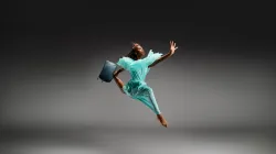 A young dancer jumping in the air wearing a blue dress and carrying a suit case.