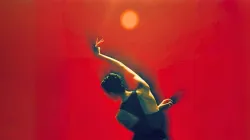 A dancer in a black costume moves in a dramatic, curved pose against a vivid red-orange background. Warm light illuminates the dancer’s outstretched arm and upper body, while a glowing circular light hovers above, creating a striking, abstract composition that evokes intensity, movement, and emotion.