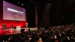 Wide shot of a large auditorium audience seated and facing a stage with a "Design 2025" screen display, a speaker standing at the podium.
