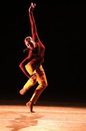 A Black woman in a burgundy shirt and tan pants dances on a stage with one arm extended in the air