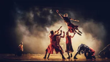 A dancer jumping in the air and four other dancers catching him. Fog covers them all.