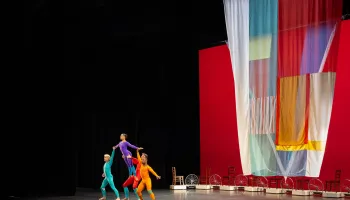 Four dancers in colorful body suite hold up one dancer as she leans forward. A colorful drape flows behind them. A row of chairs also display behind the dancers.
