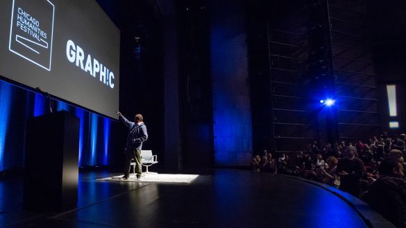 A man stands on a theater stage with blue accent lighting and a projected backdrop with white text that reads "Chicago Humanities Festival - GRAPHIC"