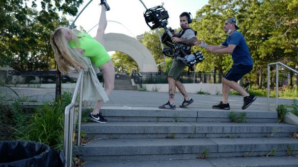 A blond woman in a green leotard and black sneakers dances with her leg extended, on steps in a park, filmed by two men in blue shirts