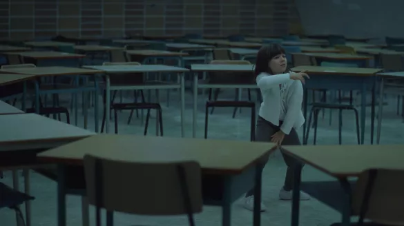 A young girl with dark hair and a white sweater dances, surrounded by desks in a classroom