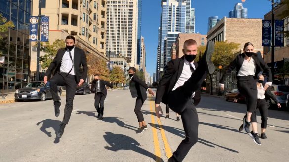 Seven Giordano Dance dancers perform in the middle of a Chicago street, tall buildings and blue sky in the background. Dancers wearing black suits with white undershirts, all caught jumping in midair.