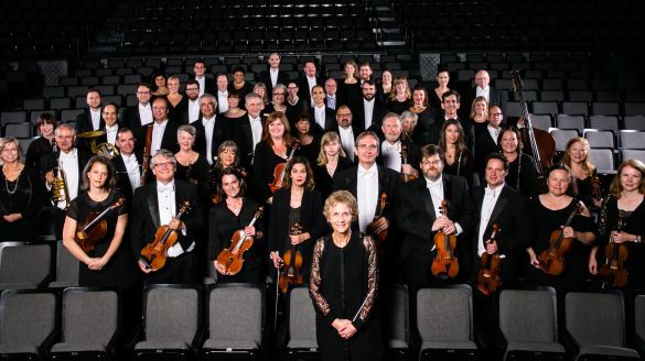 About 40 musicians in black clothing pose in the seats of an auditorium, holding various string instruments