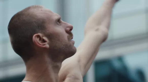 One dancer stands in profile in front of a glass building, their left arm extended diagonally out to their side. Their right arm with limb loss extends straight out from their body. Dancer's eyes are focused upwards.  