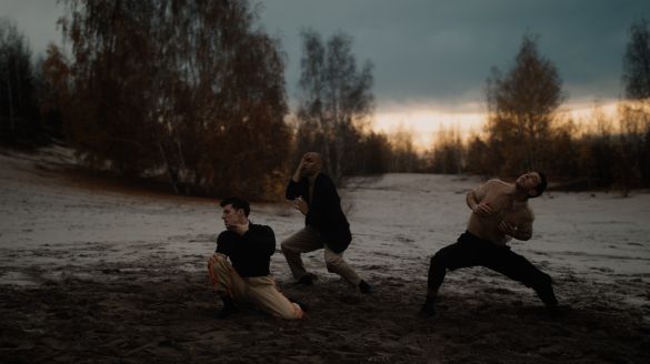 Three men in black and tan clothes dance on rocky terrain, with cloudy sky and low light on the horizon
