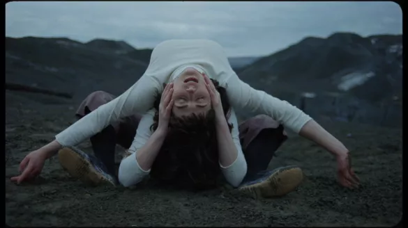 Two dancers in white shirts lie on their backs on dark green earth, one with arms extended over the other