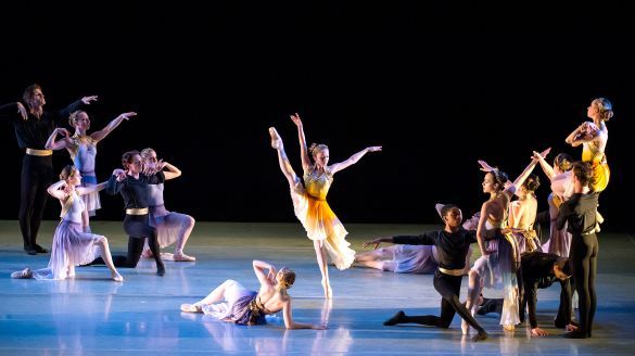 An ensemble of ballet dancers pose on a softly lit stage with blue hues, with a single female dancer at the center raising her leg high in the air