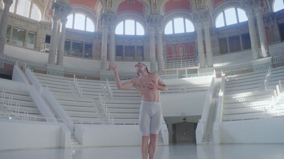 A White male dancer in white shorts poses in a sunlit, dome-ceilinged room
