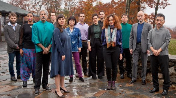 13 musicians in casual attire pose in front of trees with autumn colors