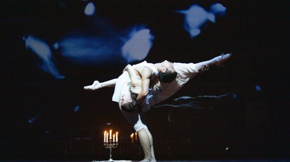 Two dancers in white pose on a dark stage illuminated by a lantern