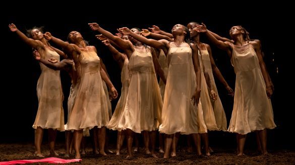 Eight Black women dancers in light dresses dance on a dark stage