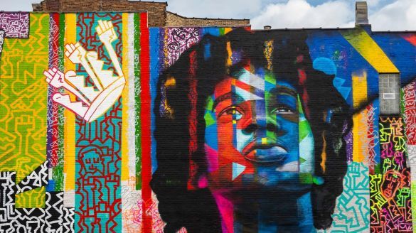 A close up of a mural painted on a brick wall, showing a young African American person looking up towards the sky. The background is multicolored with stripes and zig-zagged lines.