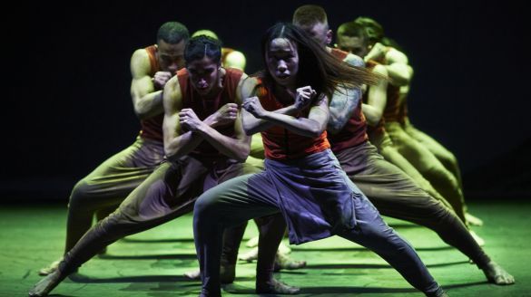 Dancers lunge with their forearms crossed on a dark stage with green lighting
