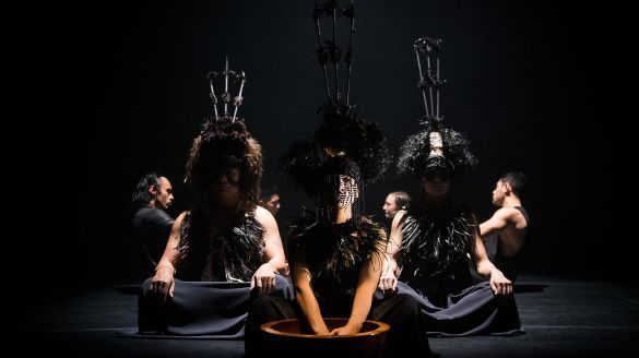 Five dancers sitting on the ground wearing black costumes with feathers and beaded ornaments.
