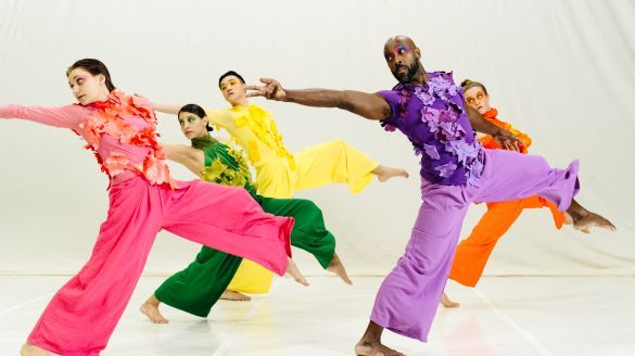 Five dancers in brightly colored, floral-appliqué costumes reach back with their arm while lunging forward with their leg. 