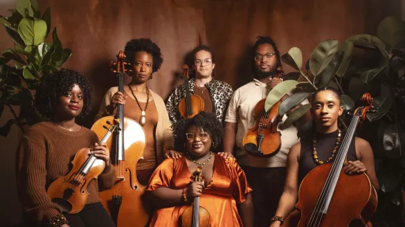 D-Composed members pose together with their instruments in front of a brown solid backdrop and plants.