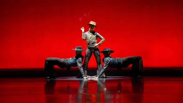 One dancer in a hat stands confidently in front of a deep red backdrop, flanked by 2 other dancers on either side.