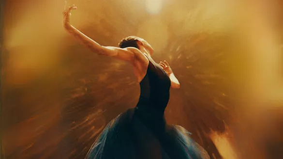 A ballerina looking away with her arms extended upward. A golden light burst and shine in the foreground.