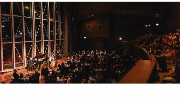 An audience seated on risers and tables watching a performance on the Pritzker stage with the Millennium Park and city skyline at night as the backdrop.