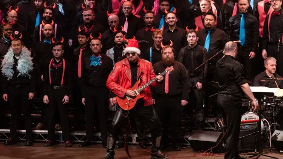 A man in a red leather jacket and Santa Claus hat plays the guitar and a choir stands behind him.