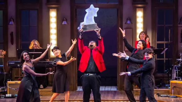 A man wearing a red blazer holds up a large star award astonished. Several others motion to him in excitment.