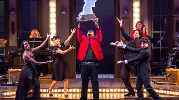 A man in all wearing a red blazer holds up a large star award astonished. Several others motion to him in excitment.