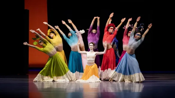 Dancers stand in the middle with their hands extended up and outward. They all wear different color skirts and shirts.