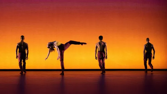 Four dancers stand in a line. One gently falls to her back lifting her leg and arms.