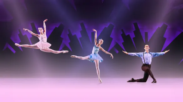 Three ballet dancers pose on stage before a purple city skyline; one leaps, one balances on pointe, and one kneels with arms extended.