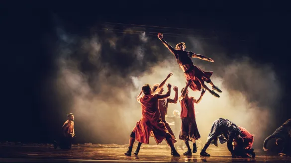 A dancer jumping in the air and four other dancers catching him. Fog covers them all.