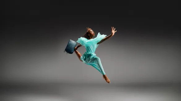 A young dancer jumping in the air wearing a blue dress and carrying a suit case.