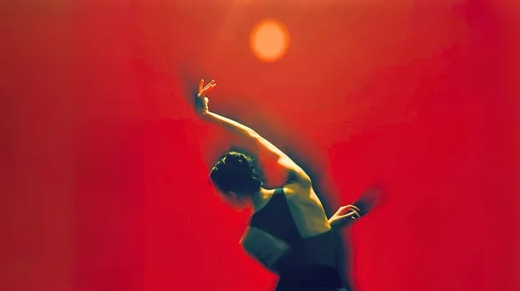 A dancer in a black costume moves in a dramatic, curved pose against a vivid red-orange background. Warm light illuminates the dancer’s outstretched arm and upper body, while a glowing circular light hovers above, creating a striking, abstract composition that evokes intensity, movement, and emotion.