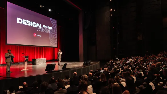 Wide shot of a large auditorium audience seated and facing a stage with a "Design 2025" screen display, a speaker standing at the podium.