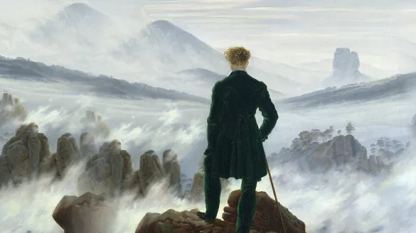with his back to the viewer, a man stands on a rock staring out at foggy mountains