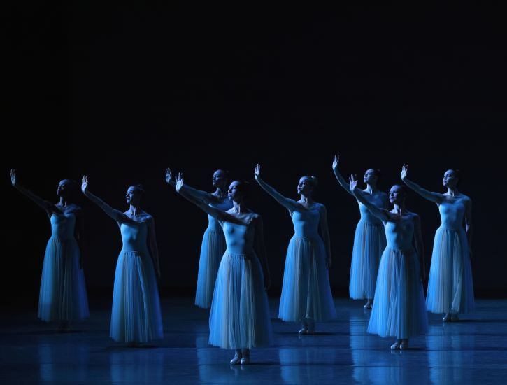 Several dancers dressed in white dresses lift their arms to stage right.