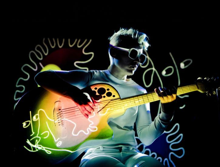 A person plays the guitar. A colorful illustration is projected on them and the guitar. 