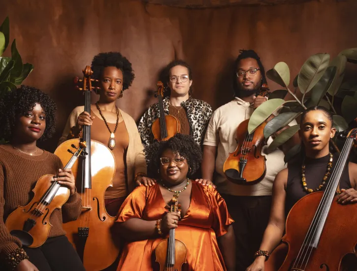 D-Composed members pose together with their instruments in front of a brown solid backdrop and plants.
