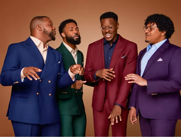 King's Return all wear different color suits. They stand in front of a solid brown backdrop as they sing.